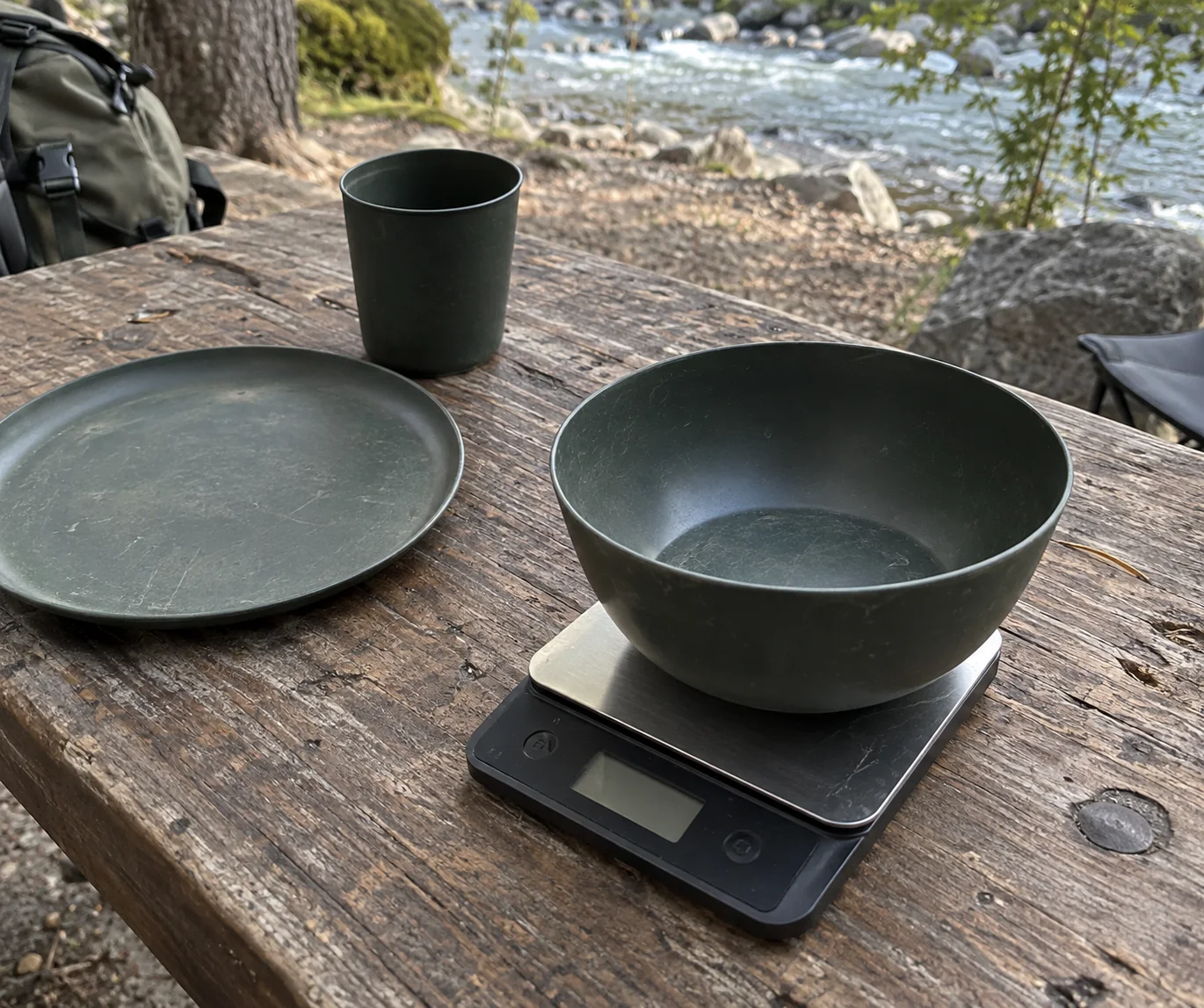Camping bowl on a scale beside a plate and cup at a riverside picnic table